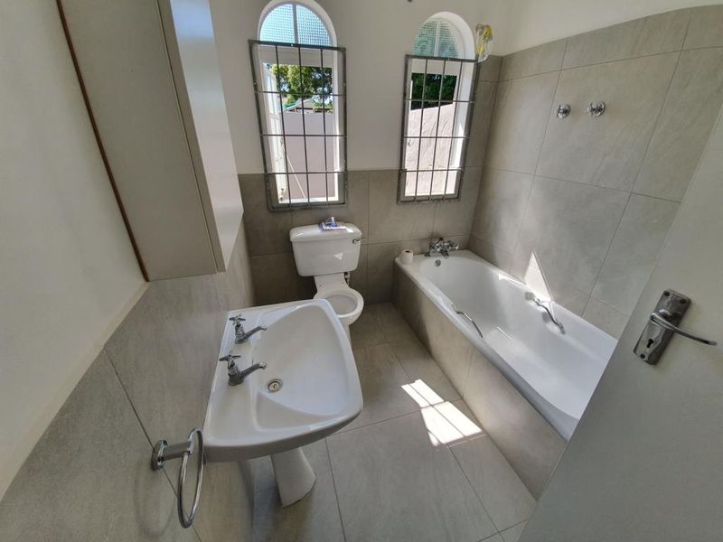 A functional bathroom featuring a pedestal basin, toilet, and bathtub with neutral wall and floor tiling and security-fitted windows.