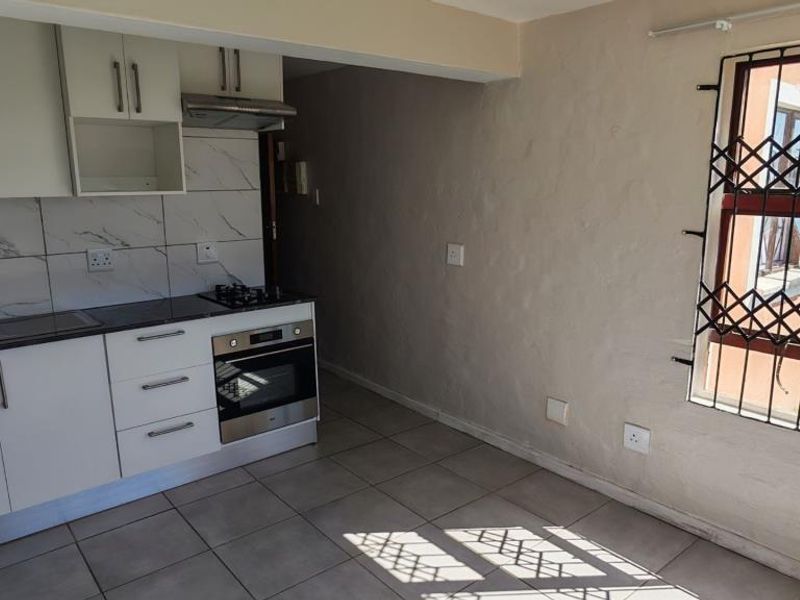 A compact kitchen featuring white cabinetry, marble-look splashback tiles, built-in oven and hob, and tiled flooring, with a security-gated window.