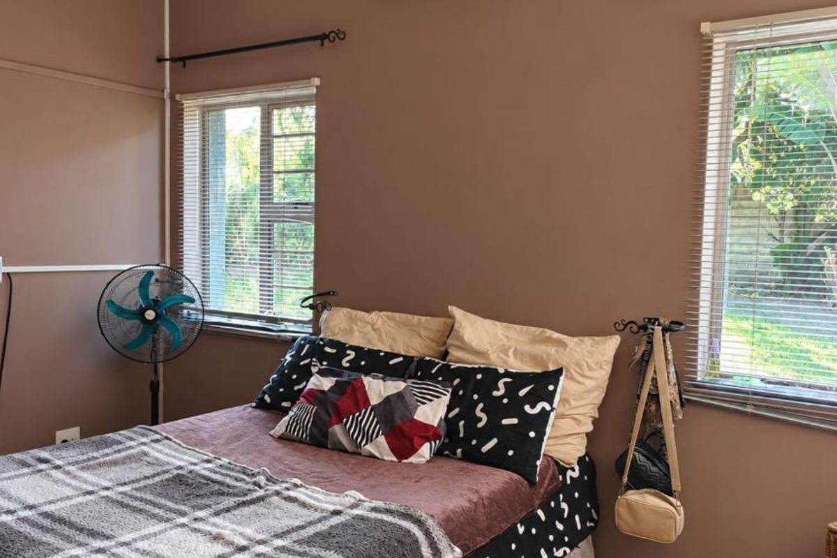 A bedroom featuring brown walls, aluminium window frames with venetian blinds, and visible electrical conduit running along the wall.