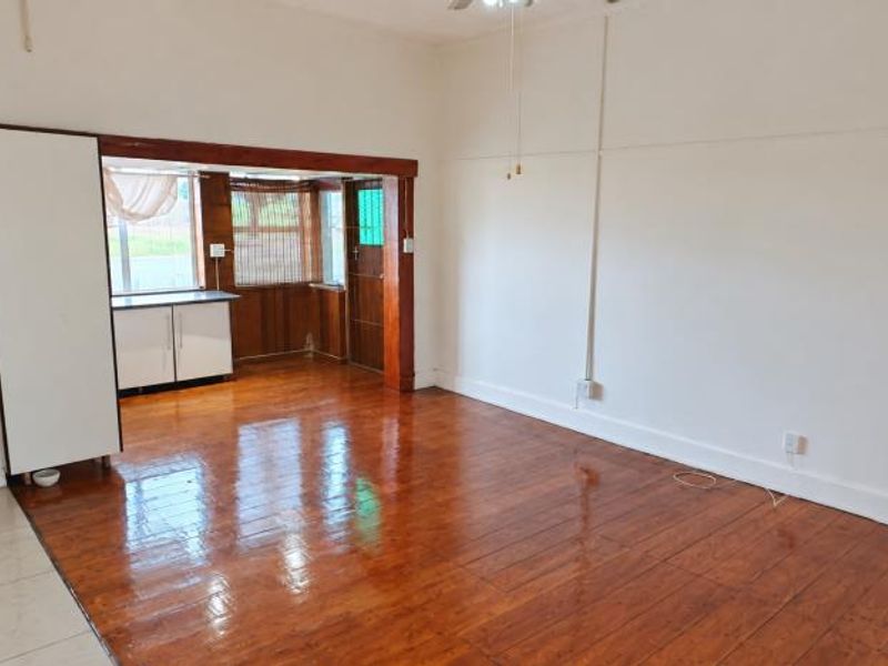 An open-plan living area featuring polished wooden floors, a ceiling fan, and a built-in cupboard, connecting to an adjacent utility or kitchen space.