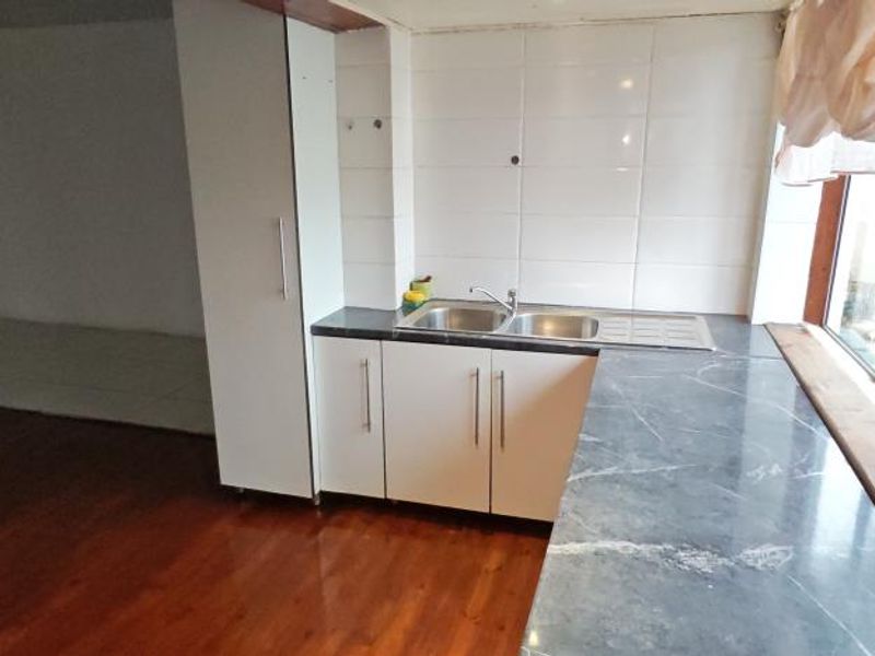 A compact kitchen area featuring a double bowl sink, tiled splashback, and dark laminate countertops, set against timber flooring with large windows.