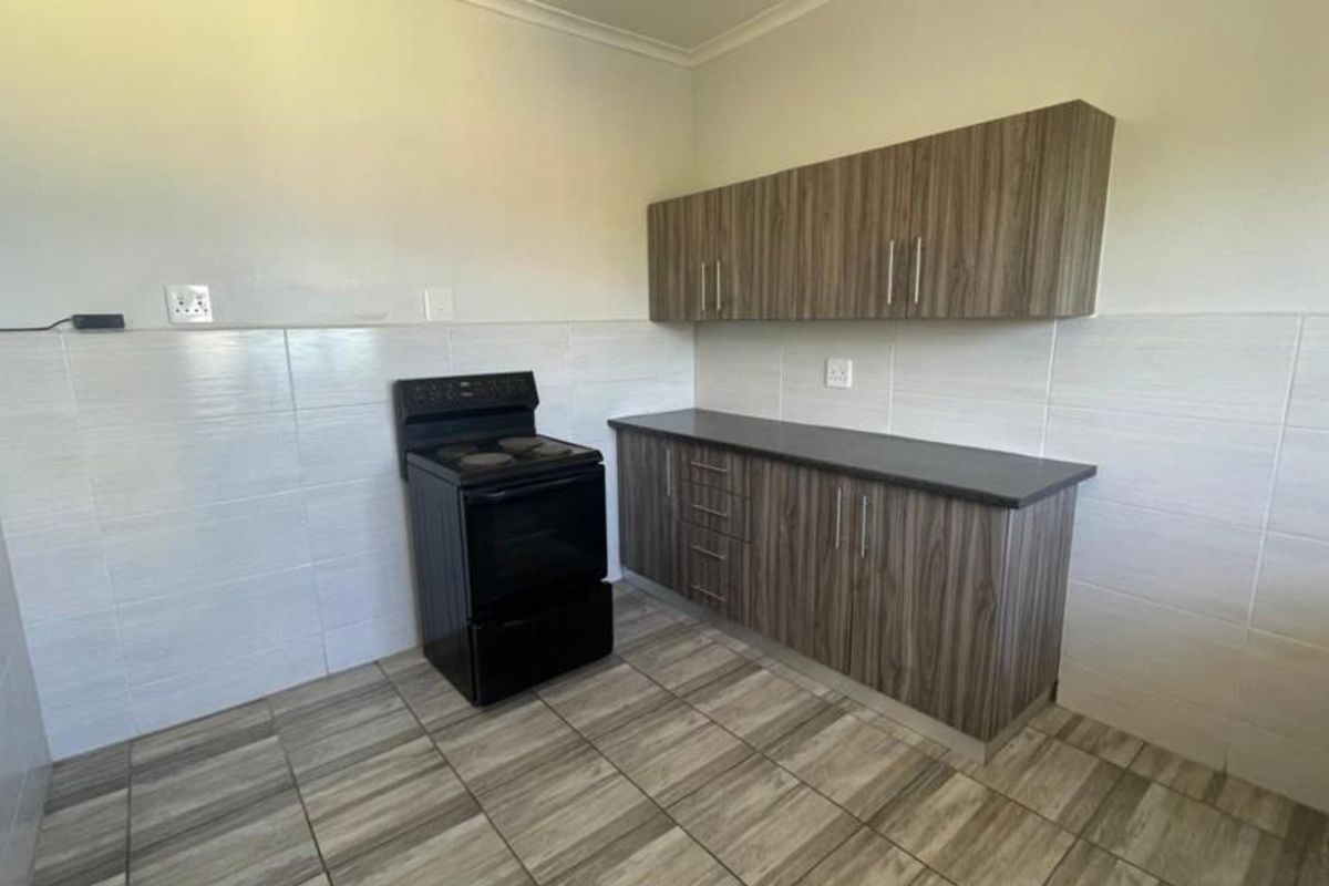 A clean, functional kitchen featuring wood-look laminate cabinetry, dark countertops, a freestanding electric stove, and neutral tiled splashbacks and flooring.