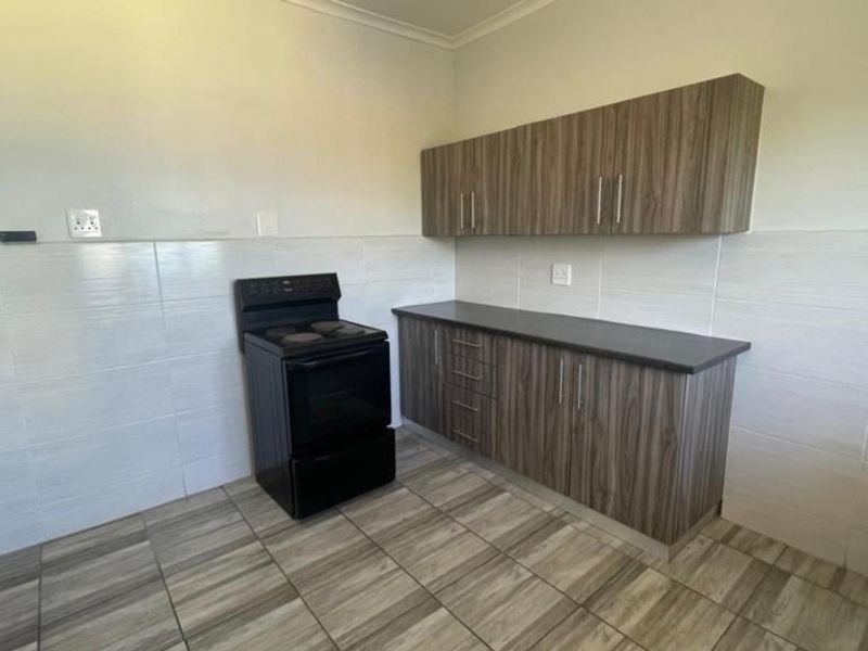A clean, functional kitchen featuring wood-look laminate cabinetry, dark countertops, a freestanding electric stove, and neutral tiled splashbacks and flooring.