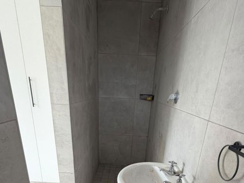 A compact bathroom featuring neutral wall tiles, a pedestal basin, and a shower area, presenting a clean and functional space.
