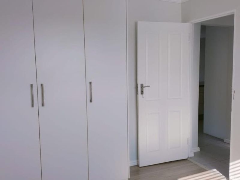 A clean, neutral bedroom featuring built-in cupboards and light-toned laminate flooring, leading into an adjacent hallway.