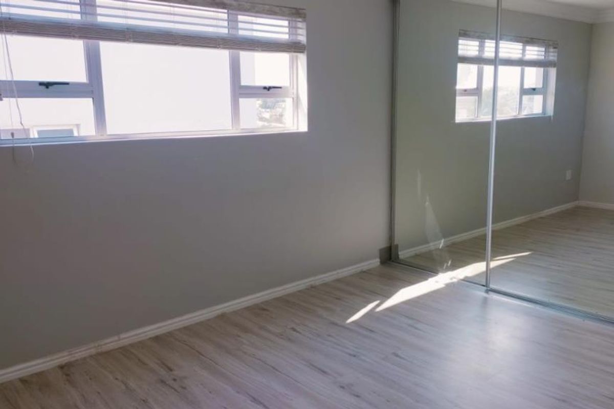 A bright, empty bedroom featuring modern laminate flooring, aluminium windows with blinds, and a large built-in mirrored wardrobe.