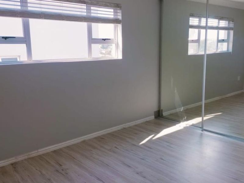 A bright, empty bedroom featuring modern laminate flooring, aluminium windows with blinds, and a large built-in mirrored wardrobe.