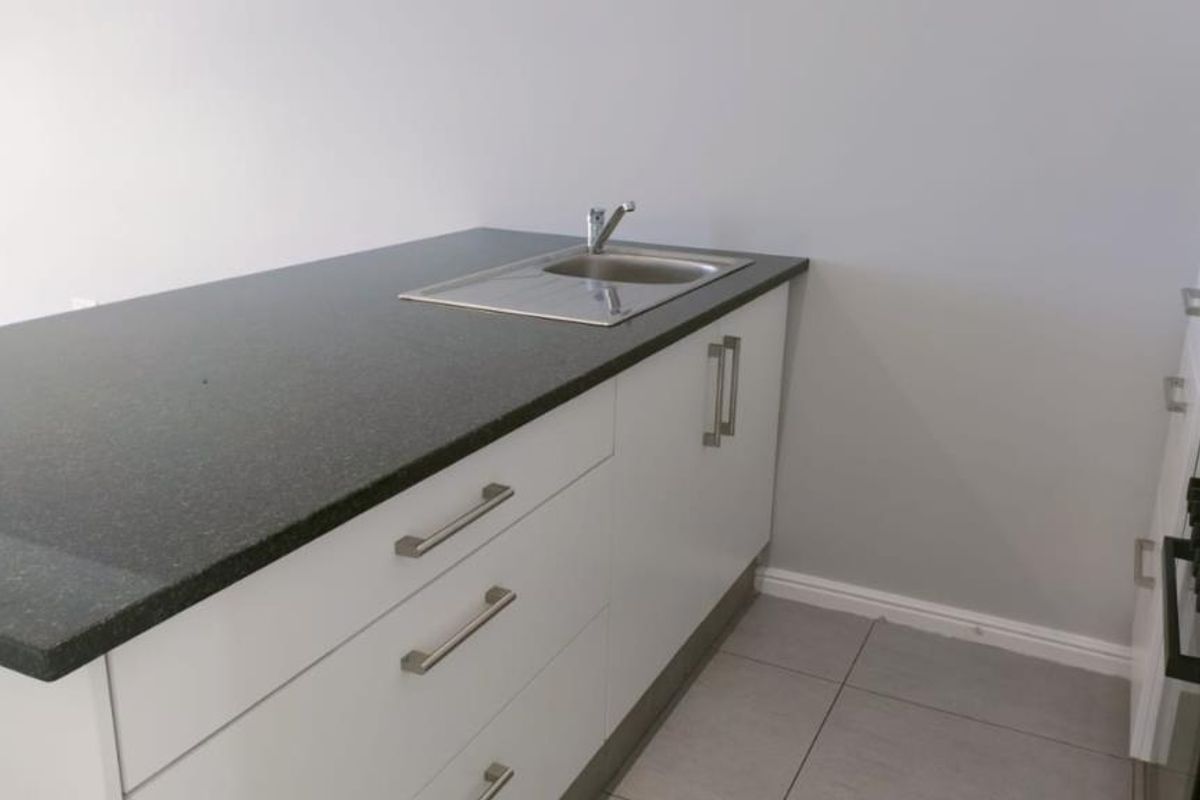 Modern kitchen area featuring white cabinetry, dark stone countertops, a stainless steel sink, and built-in oven, finished with neutral floor tiles.
