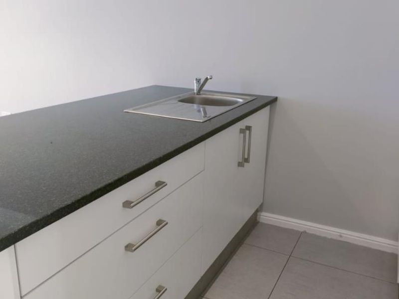 Modern kitchen area featuring white cabinetry, dark stone countertops, a stainless steel sink, and built-in oven, finished with neutral floor tiles.