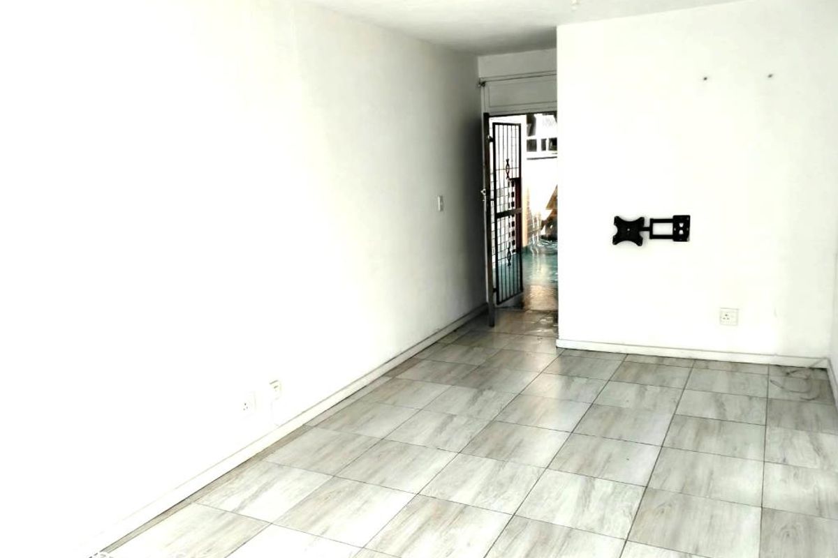 An empty living area featuring light-toned tiled flooring, a wall-mounted television bracket, and access to an exterior space through a security gate.