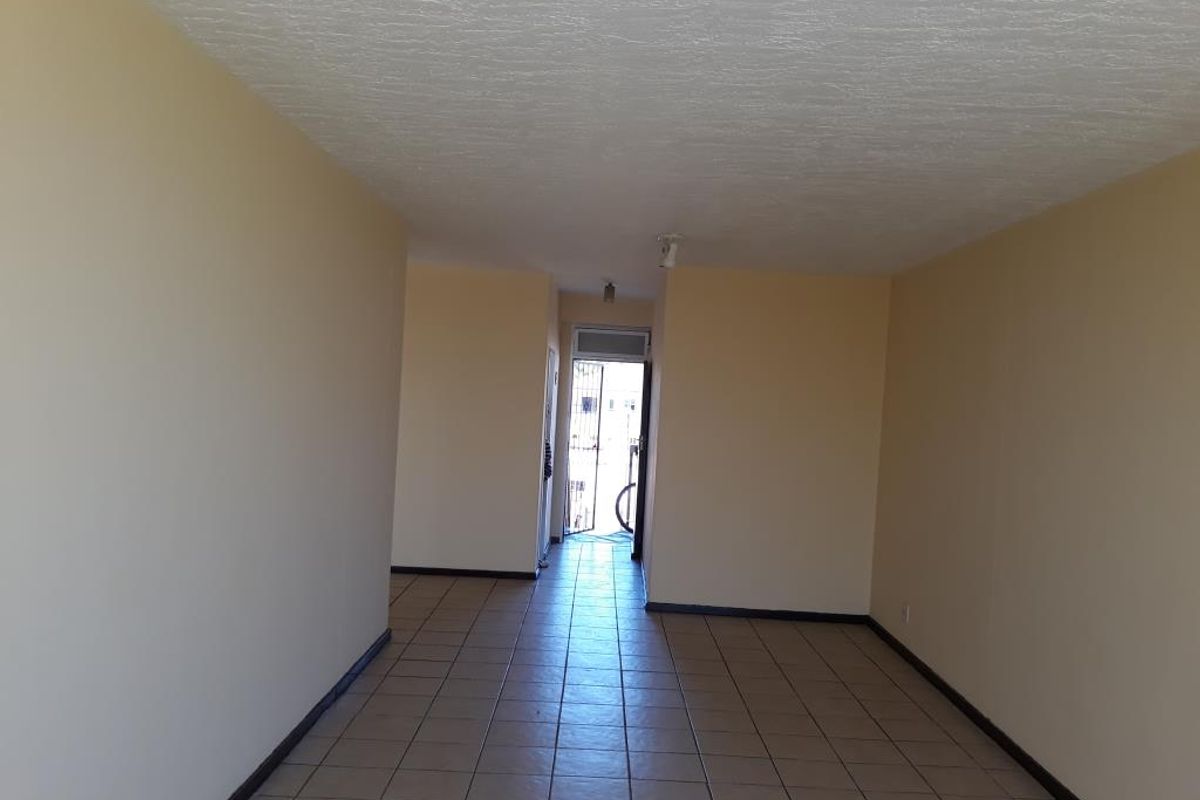 An empty, open-plan living area featuring neutral walls and tiled flooring, leading to an exterior entrance with a security gate.