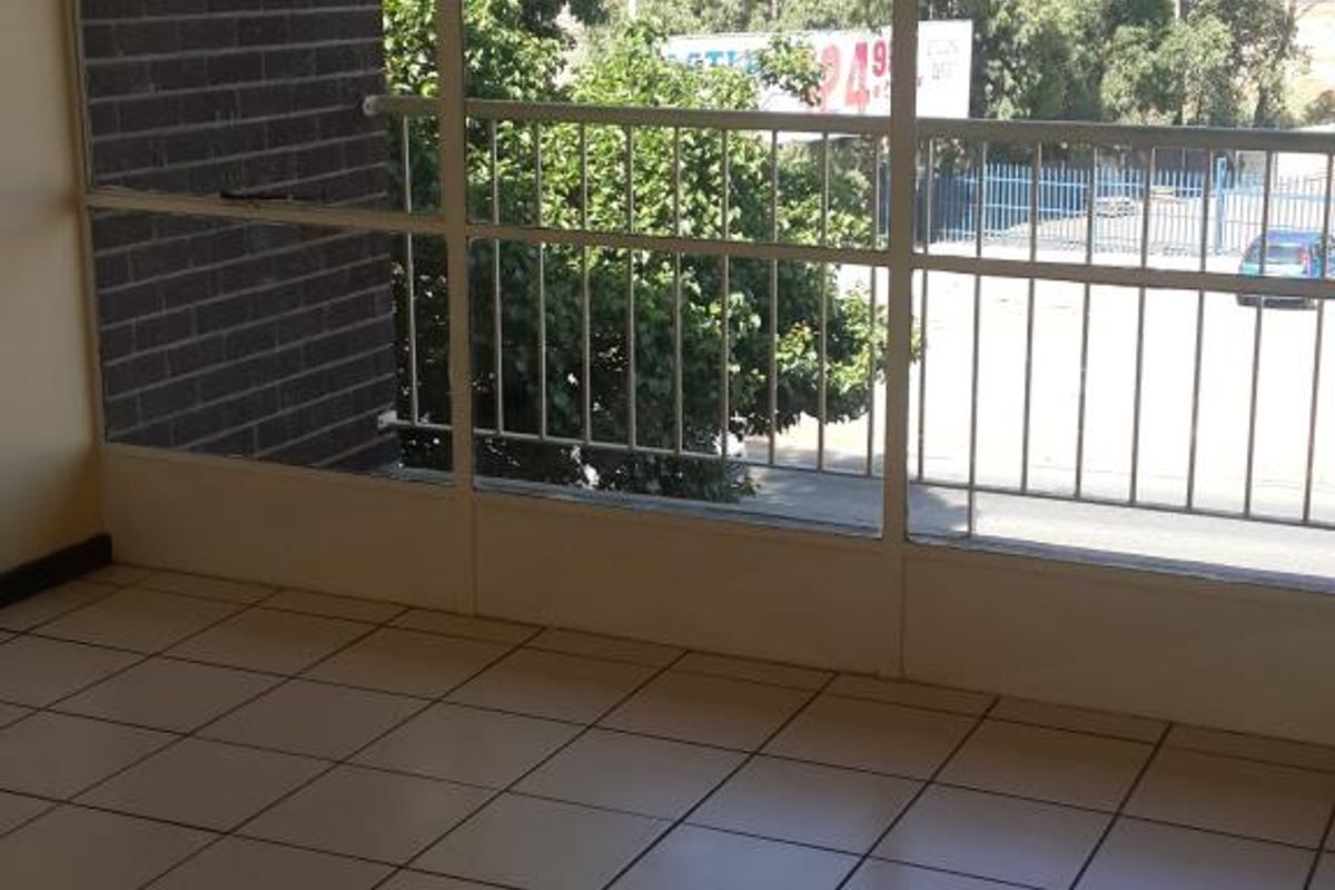An empty living area featuring large metal-framed windows, tiled flooring, and a ventilation grille, offering a view of the exterior surroundings.
