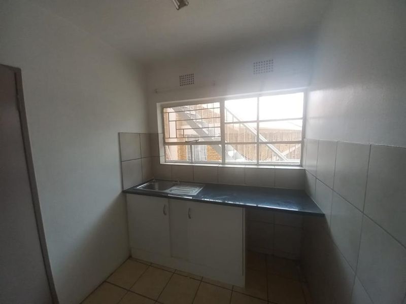 A compact, empty kitchen featuring built-in cabinetry, a stainless steel sink, tiled splashbacks, and tiled flooring with security bars on the window.