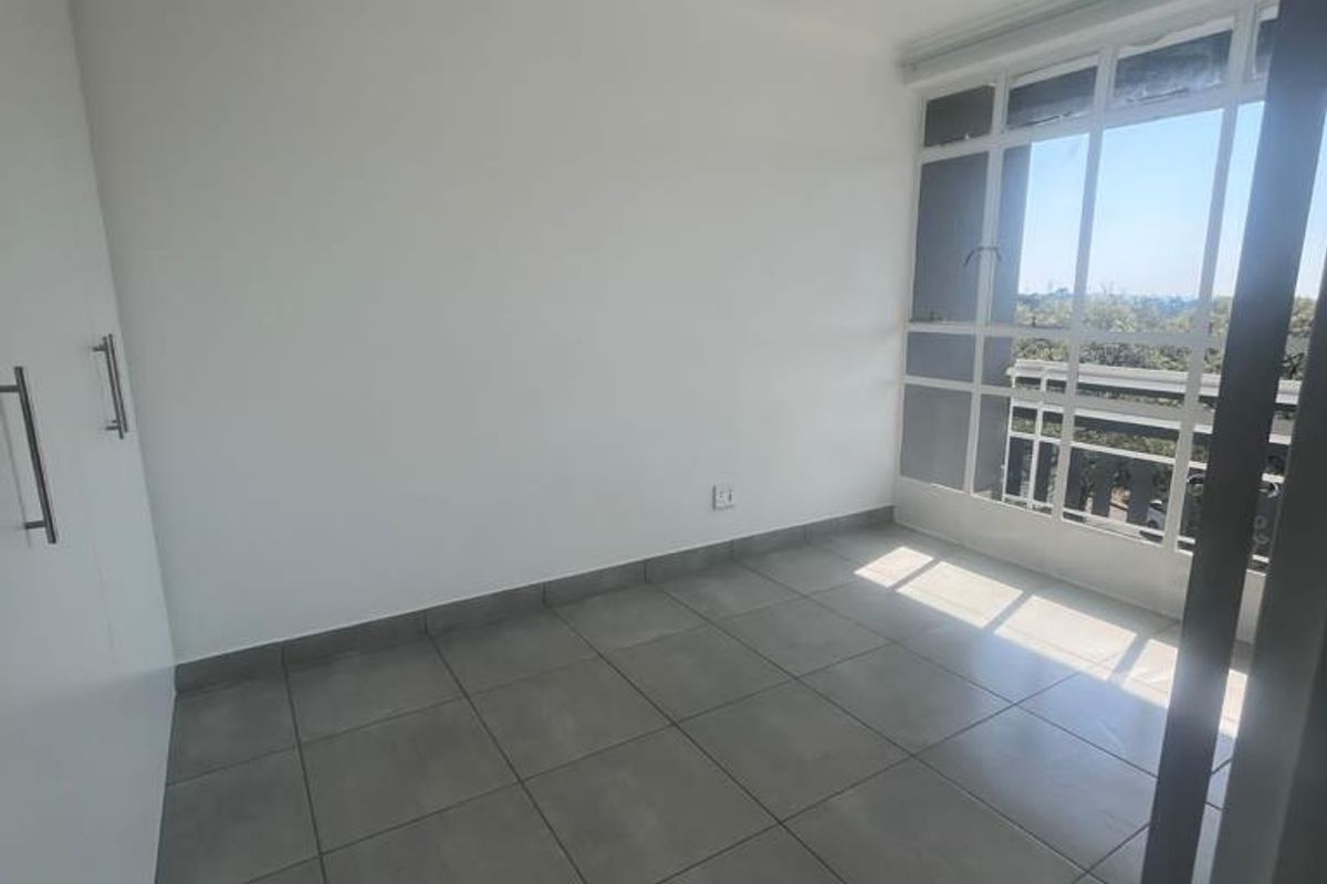 A bright, empty bedroom featuring neutral tiled flooring, built-in cupboards, and a large window providing natural light and an exterior view.