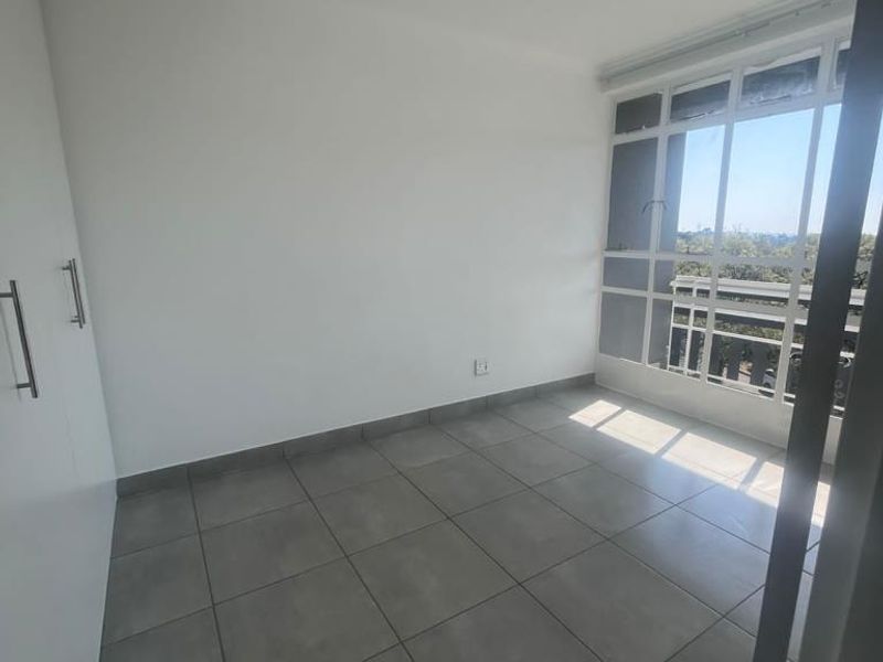 A bright, empty bedroom featuring neutral tiled flooring, built-in cupboards, and a large window providing natural light and an exterior view.