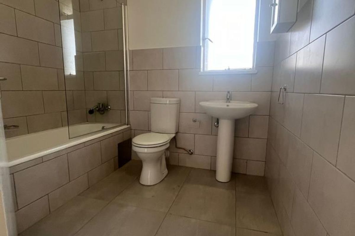 A functional bathroom featuring a bathtub with a glass screen, pedestal sink, toilet, and neutral wall and floor tiling.