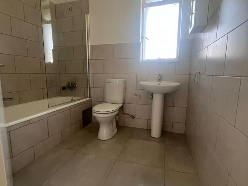 A functional bathroom featuring a bathtub with a glass screen, pedestal sink, toilet, and neutral wall and floor tiling.