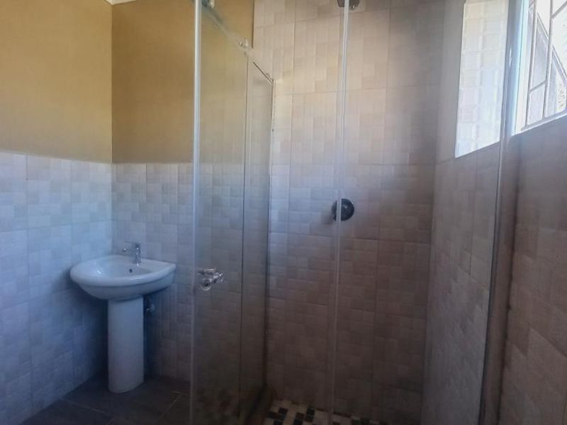 A clean, functional bathroom featuring a glass-enclosed shower, pedestal basin, and neutral wall tiling with a decorative floor pattern.