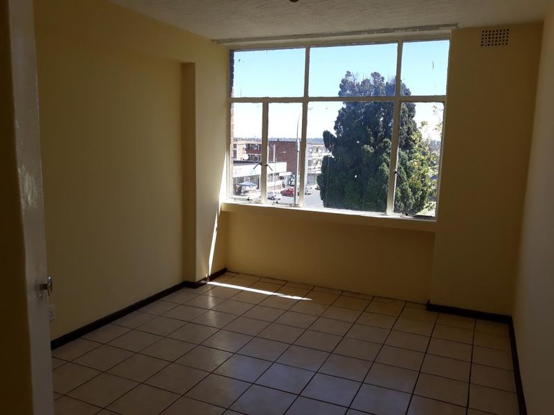 An empty living room featuring large windows with natural light, tiled flooring, and neutral wall finishes, offering a blank canvas for interior design.
