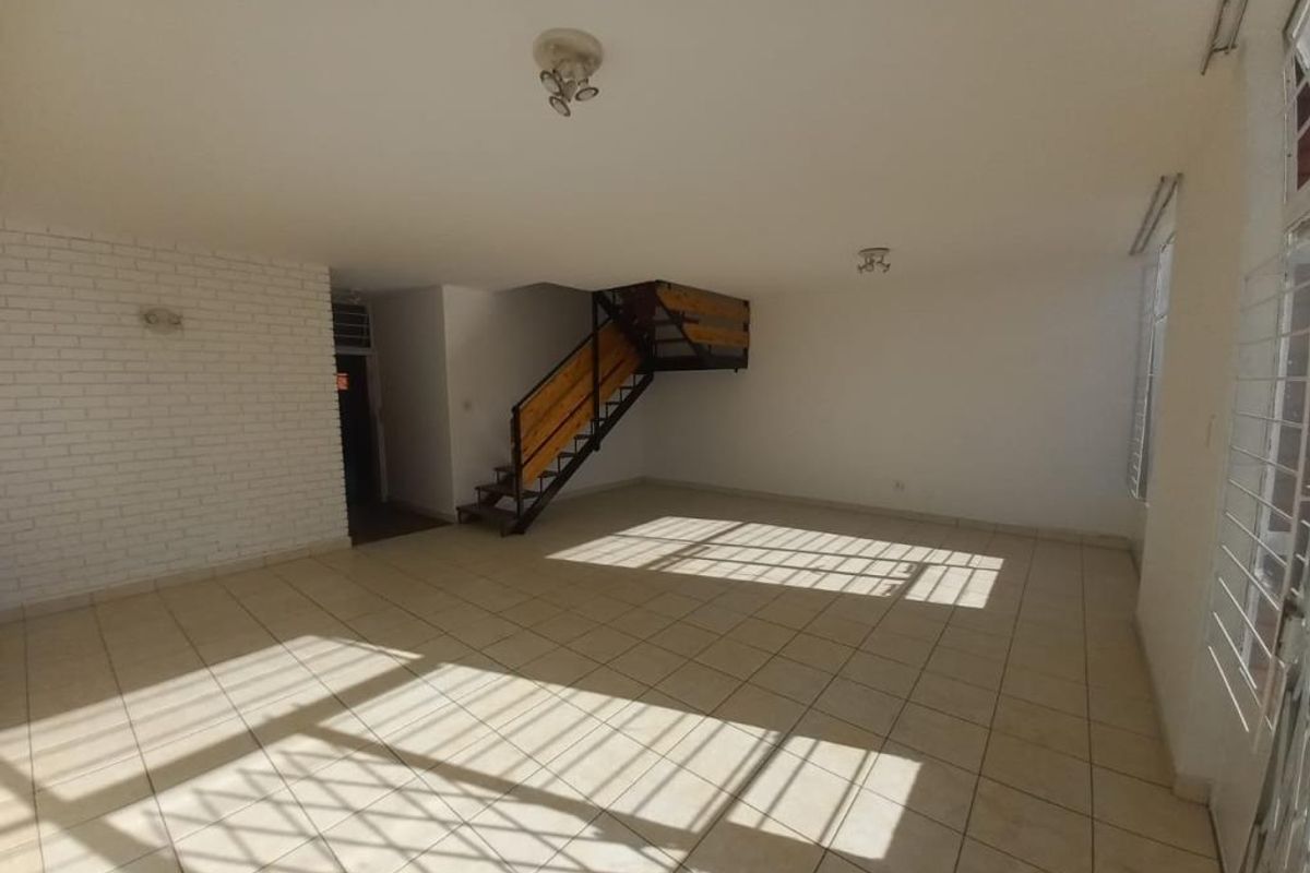 Spacious open-plan living area featuring tiled flooring, a distinctive staircase, and an exposed brick feature wall, offering a bright and versatile layout.