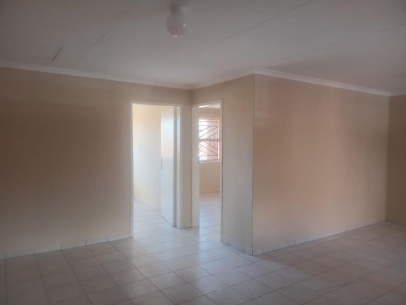 An empty, neutrally painted living area featuring tiled flooring and access to adjacent rooms through standard doorways.
