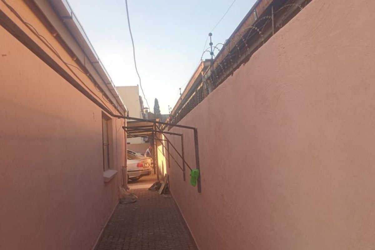 A narrow exterior side passage featuring paved flooring, high boundary walls with security razor wire, and wall-mounted metal support brackets.