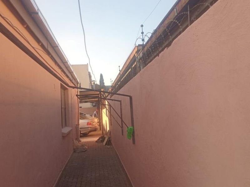 A narrow exterior side passage featuring paved flooring, high boundary walls with security razor wire, and wall-mounted metal support brackets.