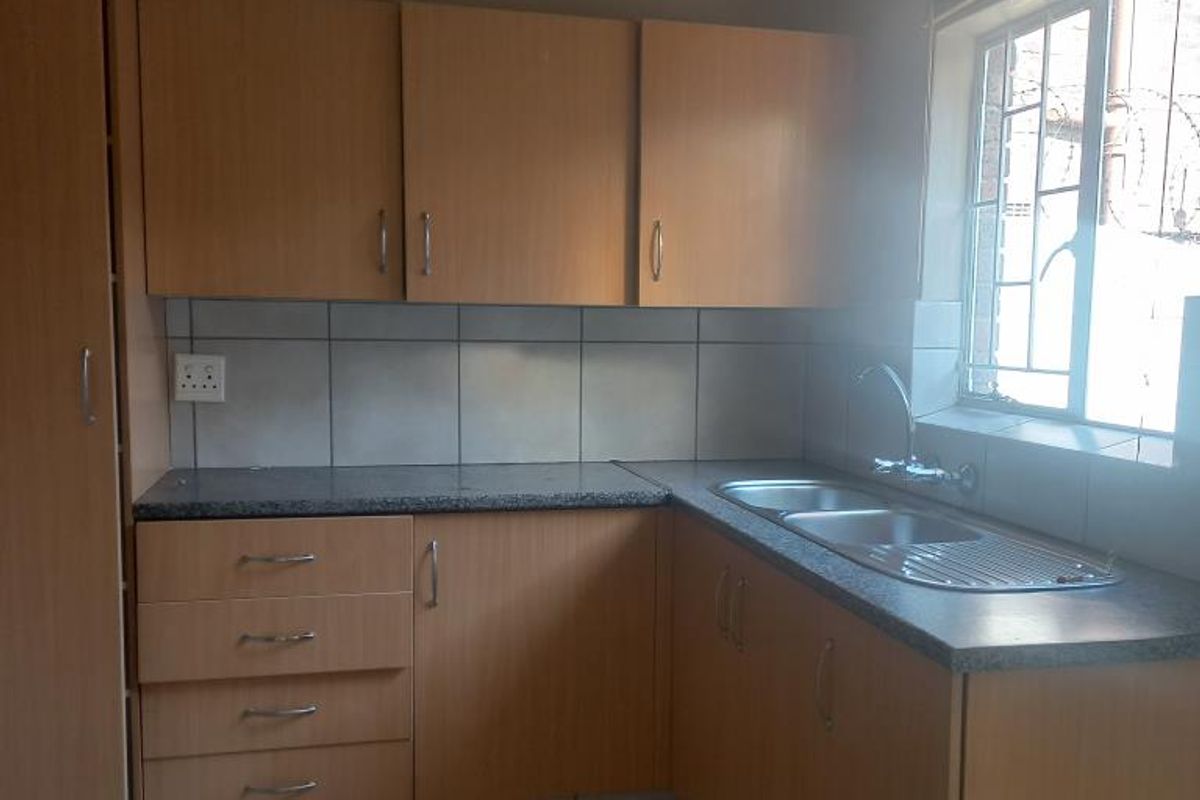 A functional kitchen featuring light wood-look laminate cabinetry, granite-style countertops, a double stainless steel sink, and neutral ceramic floor tiles.