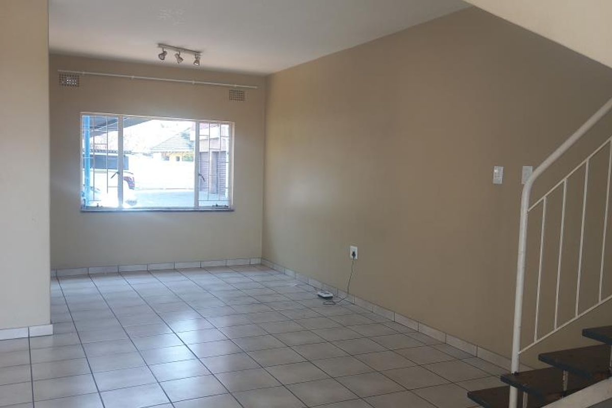 An open-plan living area featuring neutral tiled flooring, a staircase with a metal railing, and a window with security bars.
