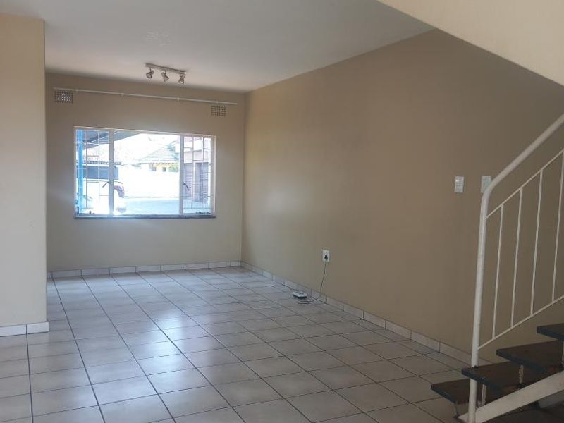 An open-plan living area featuring neutral tiled flooring, a staircase with a metal railing, and a window with security bars.
