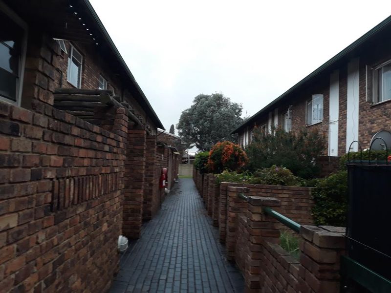 A narrow paved walkway between two brick apartment blocks, featuring low brick boundary walls and established garden greenery.