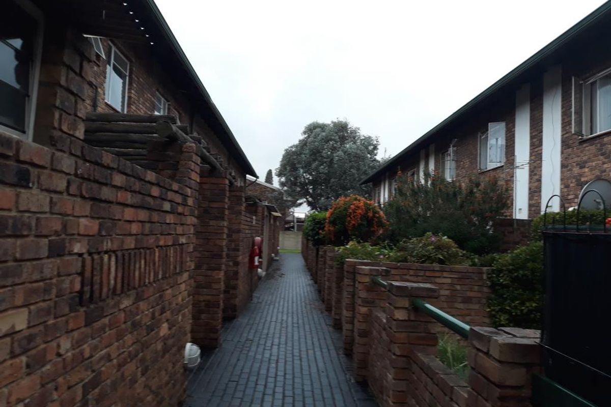 A narrow paved walkway between two brick apartment blocks, featuring low brick boundary walls and established garden greenery.