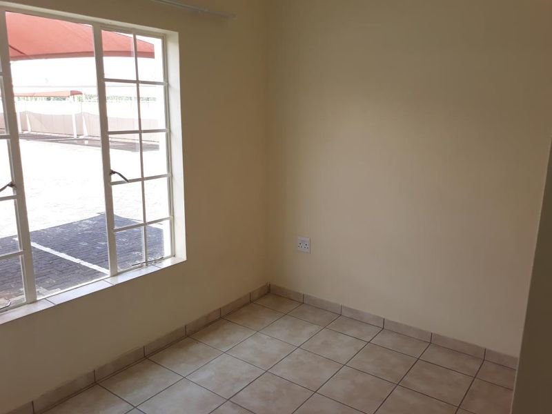 A bright, empty bedroom featuring neutral wall tones, tiled flooring, and a large window providing natural light and a view of the exterior parking area.