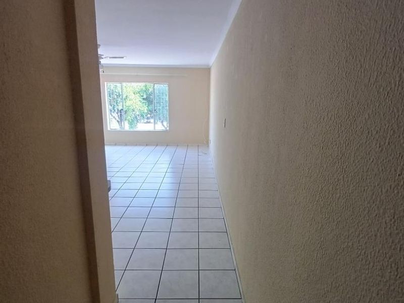A vacant hallway leading into a bright, tiled living area with a large window and ceiling fan.