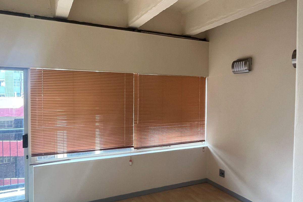 A bright, empty living area featuring laminate flooring, large windows with fitted blinds, and exposed structural ceiling beams.