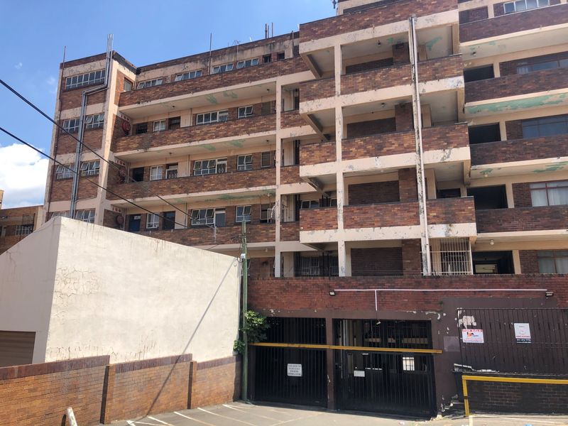 Exterior view of a multi-story brick apartment building featuring balconies, a ground-level parking area, and secure gated access.
