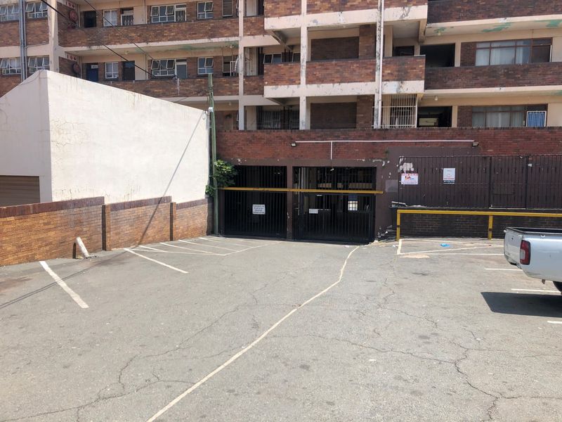 Exterior view of the flat complex featuring a paved parking area, secure gated access, and multi-story brick facade with visible signs of wear.