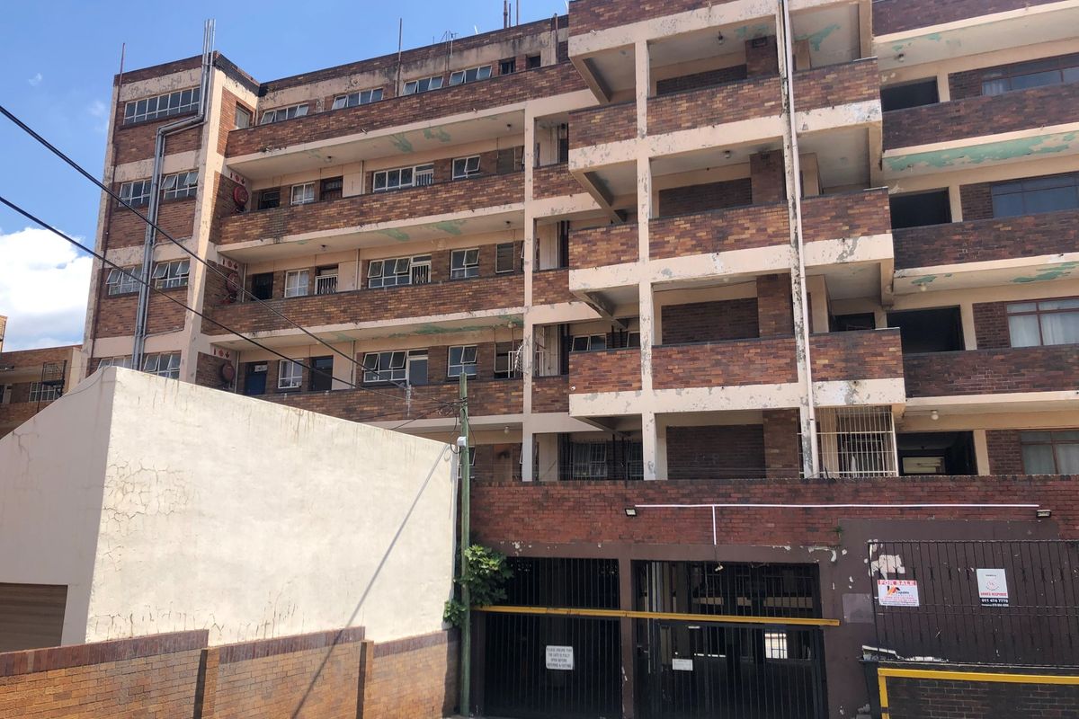 Exterior view of a multi-story brick apartment building featuring balconies, a ground-level parking area, and secure gated access.