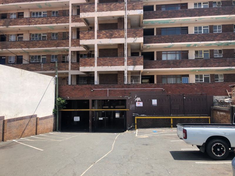 Front exterior view of a multi-story brick apartment building featuring a ground-level parking area with security gates and visible exterior wear.