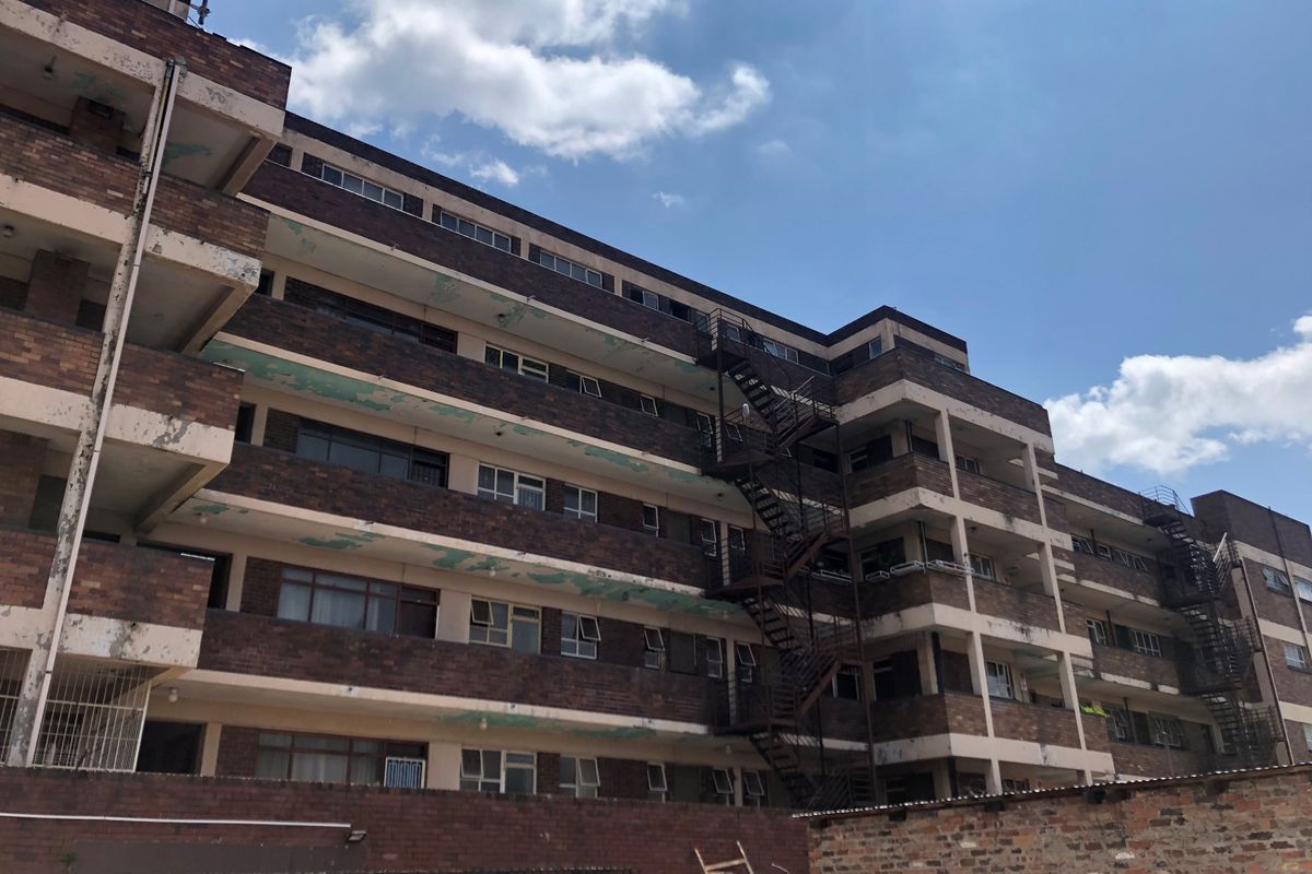 Exterior view of a multi-story brick apartment building featuring external fire escape staircases and balcony structures showing signs of weathering.