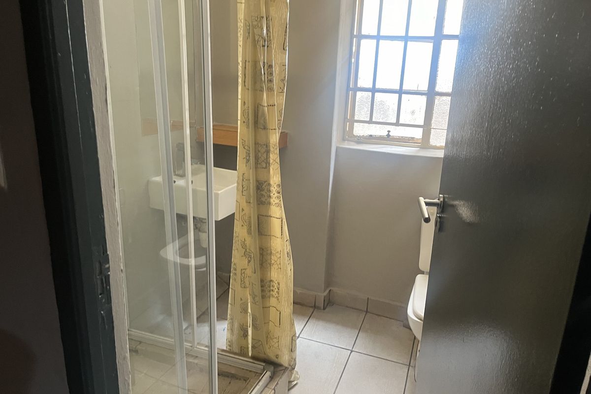 A compact bathroom featuring a shower enclosure, wall-mounted basin, and toilet, with tiled flooring and a security-barred window.