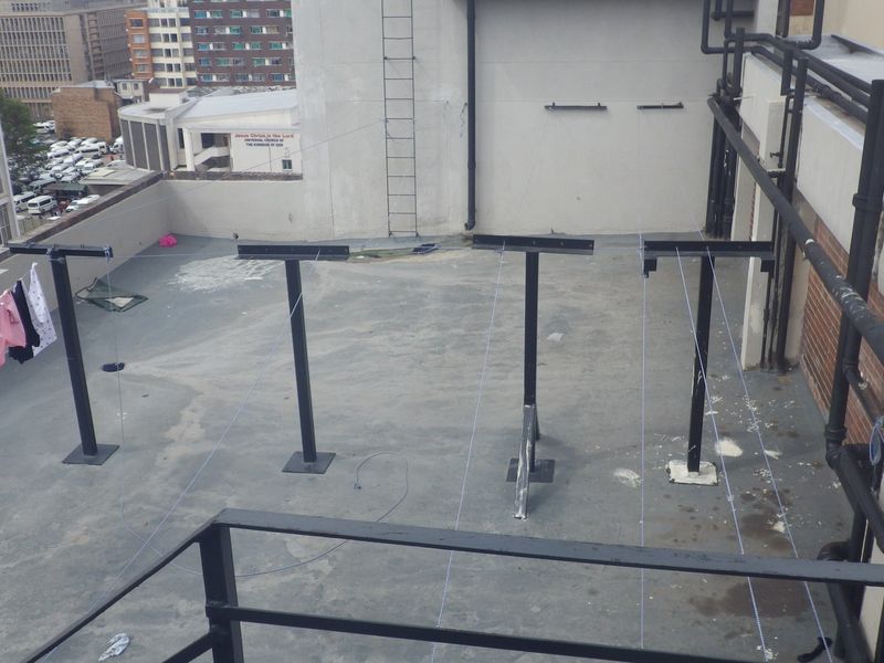 An open-air rooftop balcony area featuring metal washing line infrastructure, exposed plumbing, and concrete flooring with urban city views.
