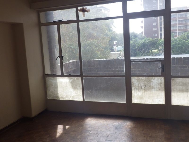 An empty living area featuring original parquet flooring and large windows providing natural light and an urban outlook.