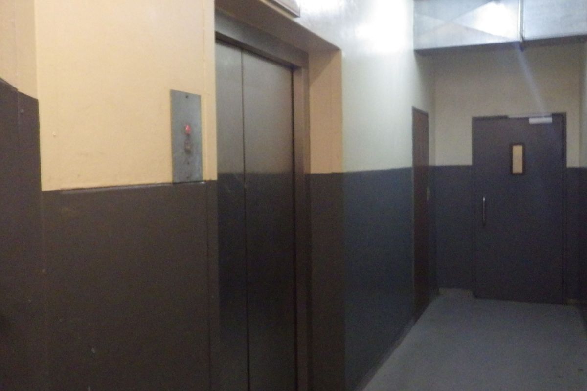 A utilitarian hallway featuring a metal elevator door, a fire-rated door, and exposed ventilation ducting, showing signs of wear on the walls.