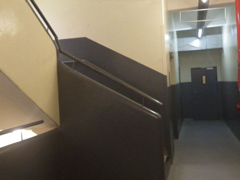 A utilitarian hallway and stairwell area featuring industrial fire safety equipment, including a hose reel and exposed piping.