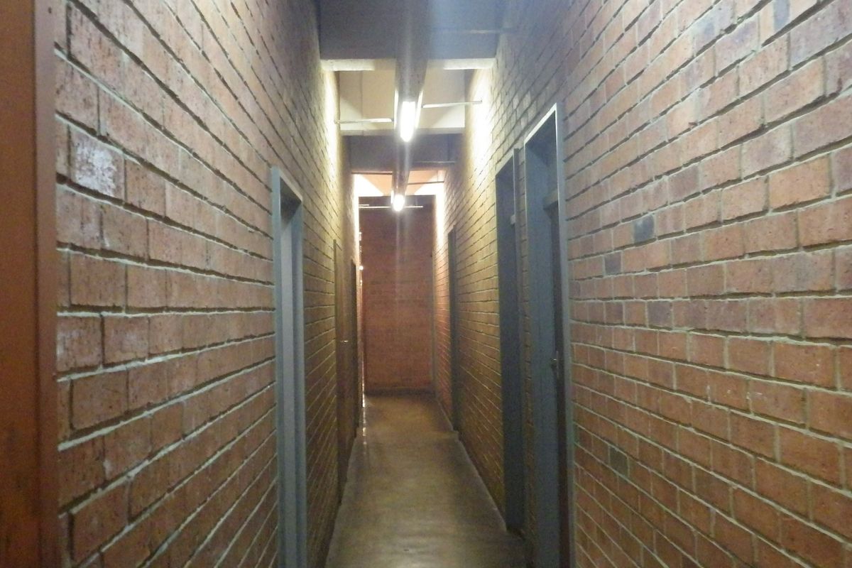 A narrow hallway featuring exposed brick walls, concrete flooring, and overhead fluorescent lighting, providing access to multiple rooms.