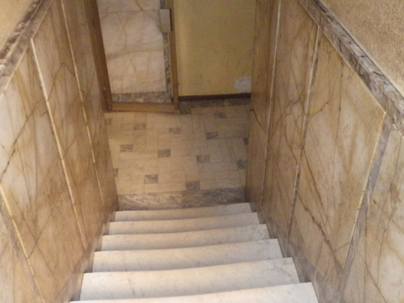 A staircase featuring marble wall cladding and marble steps, leading to a tiled landing area in a dated interior hallway.