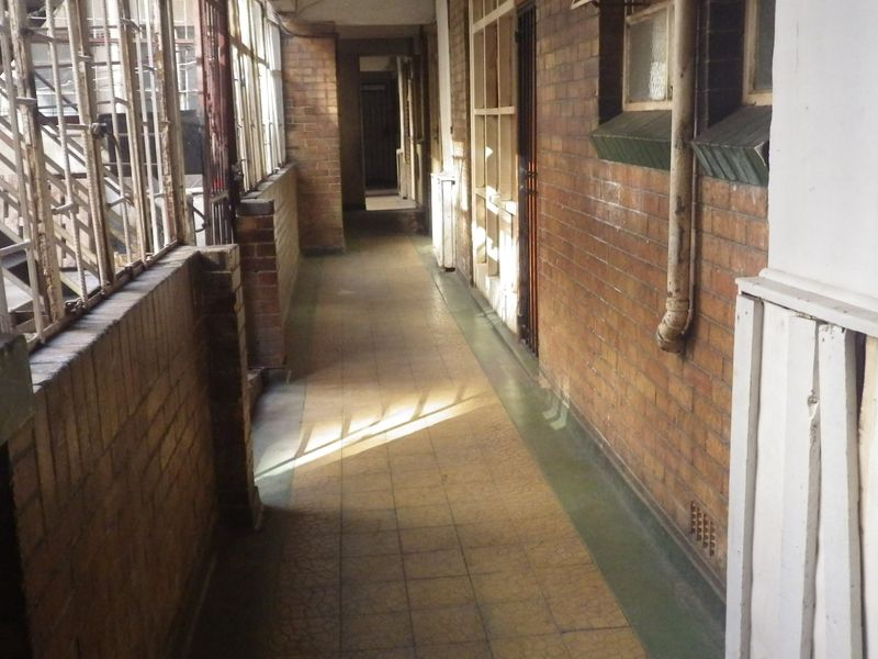 An open-air apartment building hallway featuring exposed brick walls, tiled flooring, and metal security gates, showing signs of age and wear.