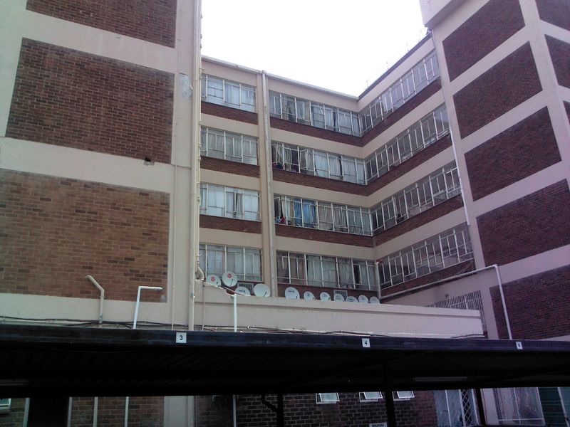 Exterior view of a multi-story brick apartment building featuring steel-framed windows, satellite dishes, and a ground-level carport structure.