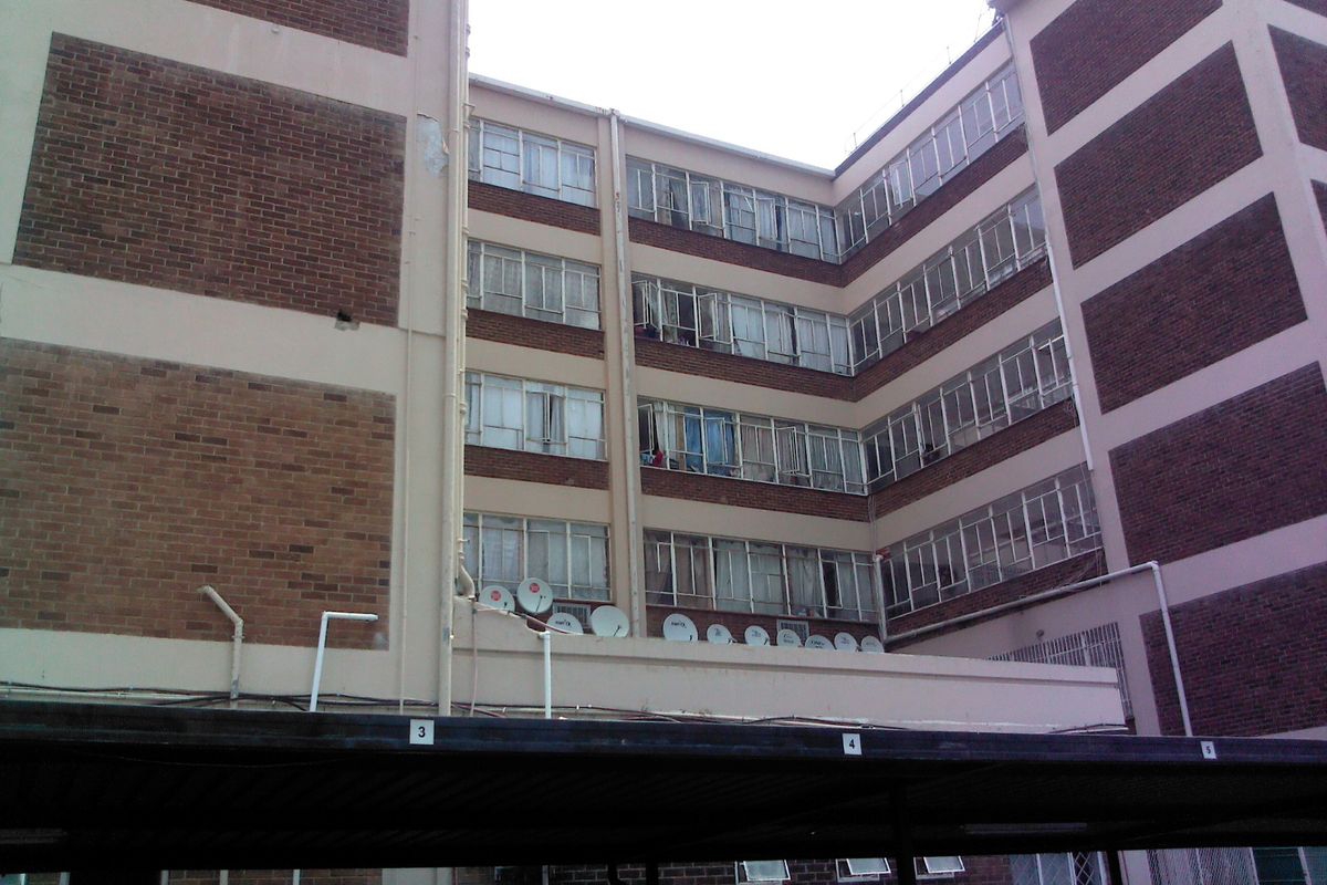 Exterior view of a multi-story brick apartment building featuring metal frame windows, satellite dishes, and a ground-level carport structure.